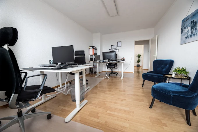 Helles Büro mit zwei Schreibtischen, Stühlen und einer Pflanze in der Ecke.