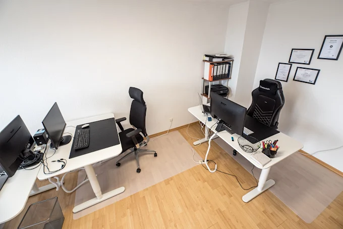 Büro mit zwei Schreibtischen, Computern, Bürostühlen und einem Regal.